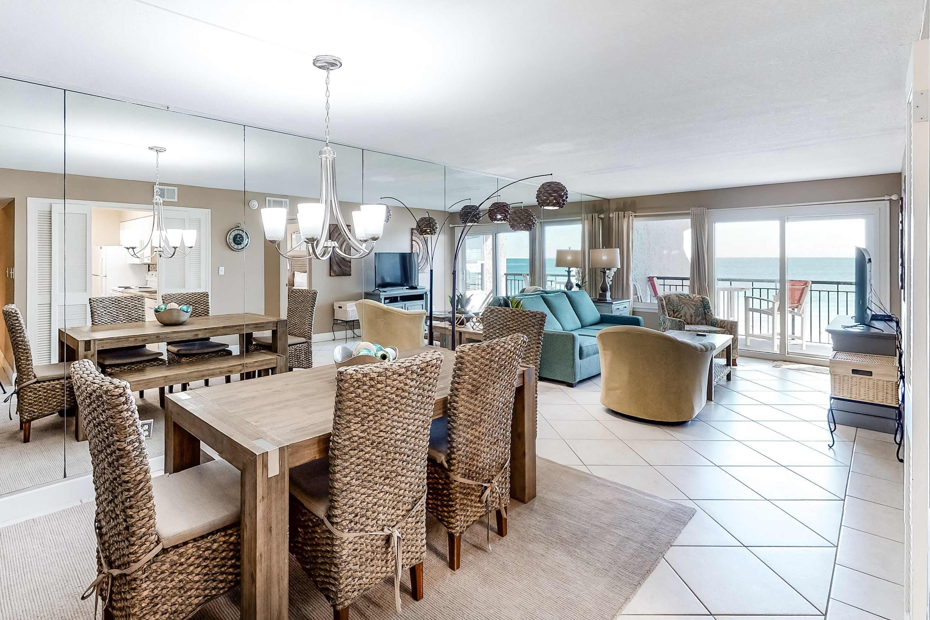 A spacious living area with a wooden dining table, wicker chairs, a sofa, and a balcony with a view of the sea.