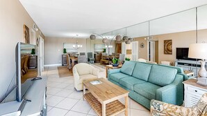 A modern living room with a sofa, coffee table, and TV. There is a dining area in the background with chairs and a table. - Updated, beachfront condo with beach setup - quick drive to entertainment (Fort Walton Beach)