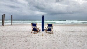 Zwei Liegestühle und ein blauer Sonnenschirm an einem sandigen Strand, im Hintergrund das Meer.