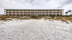 Ein mehrstöckiges Hotel mit flachem Dach und Balkonen, an einem Sandstrand mit spärlicher Vegetation gelegen.