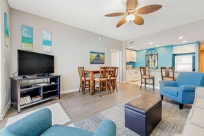 A modern living room with a flat-screen TV, wooden dining table, and blue upholstered chairs. - Beachfront condo with balcony, pool & 2 complimentary beach chairs (Panama City Beach)