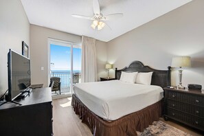 Habitación de hotel con una cama grande, un escritorio con computadora, televisión, ventilador de techo y mesita de noche con lámparas, y vista al océano a través de una puerta corrediza.