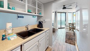 Een moderne keuken met een spoelbak, aanrecht en opbergkasten. Op de muur hangt een bordje met daarop 'NAAR HET STRAND'. De ruimte heeft uitzicht op de oceaan door grote ramen.