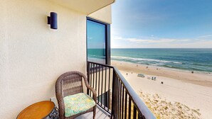 Een balkon met een rieten stoel en een klein tafeltje, met uitzicht op een strand met mensen en een heldere lucht.