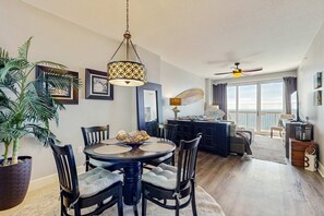 A dining area with a round table, chairs, and a hanging light fixture. There is a large potted plant and a view of the balcony through the window. - Gorgeous beachfront condo, shared pool and beach access (Panama City Beach)
