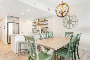 A modern kitchen with white cabinets, a stainless steel refrigerator, and a wooden dining table with green chairs. - Pristine, waterfront condo with outdoor pool, hot tub & fitness room (Panama City Beach)