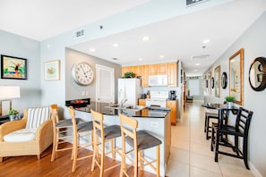 A modern kitchen with wooden cabinets, a central island with bar stools, a dining table with chairs, and a large wall clock. - Colorful, beachfront condo with Gulf view, pool, hot tub, and lazy river (Panama City Beach)