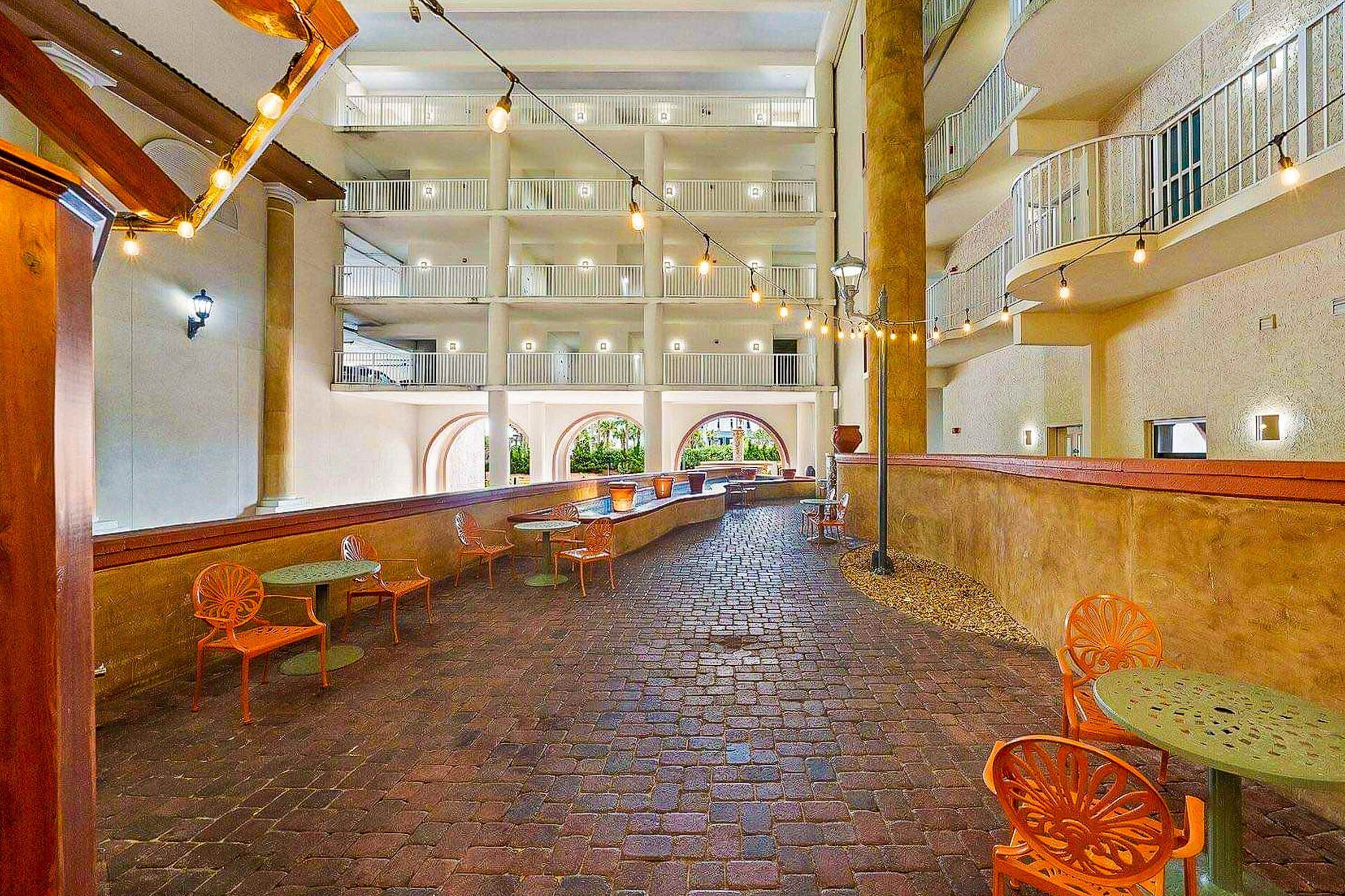 Un atrium à plusieurs niveaux avec un sol en briques, un espace extérieur aménagé avec des chaises orange et des guirlandes lumineuses.