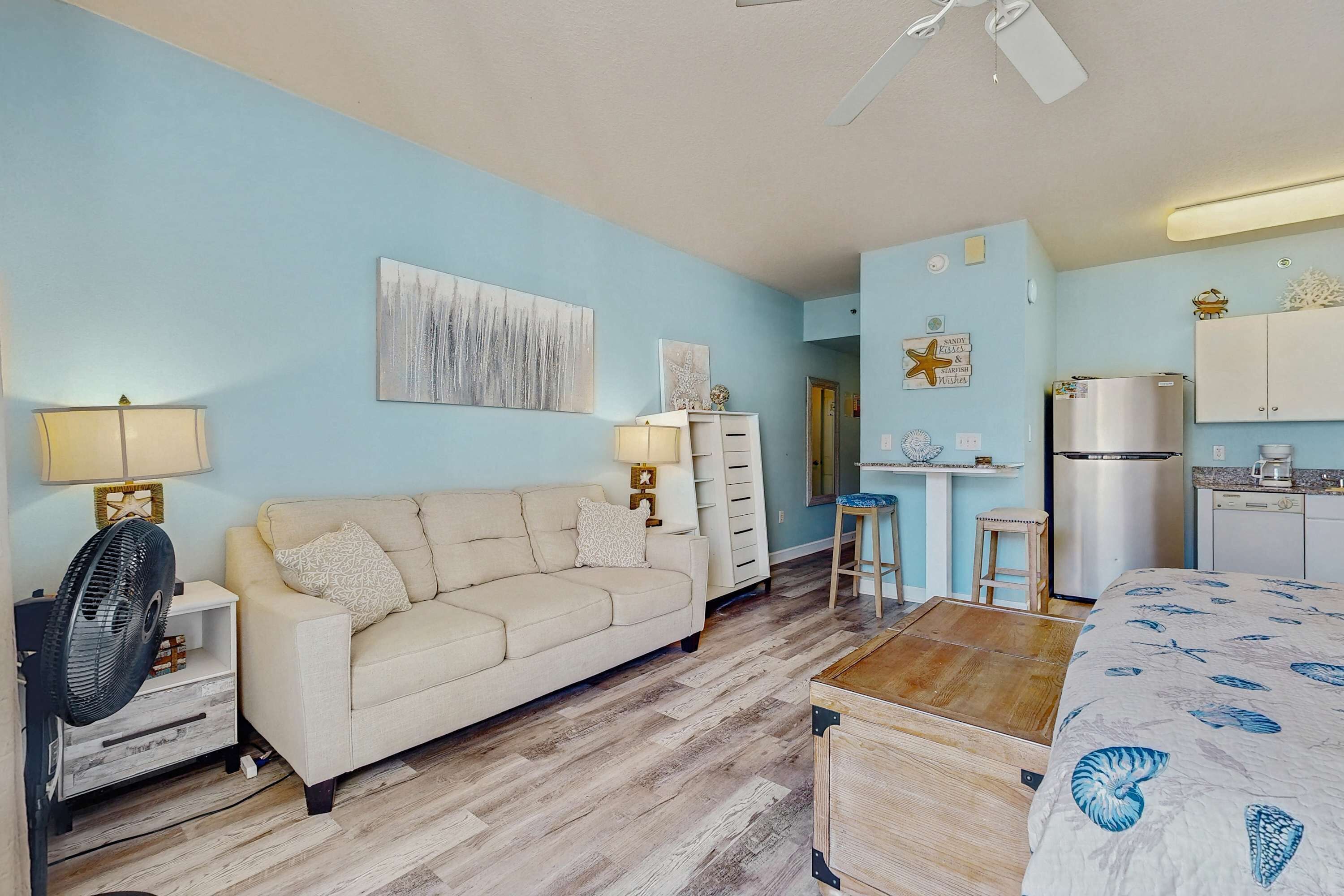 A compact living space with a sofa, a fan, a small table, a kitchenette, and a ceiling fan.