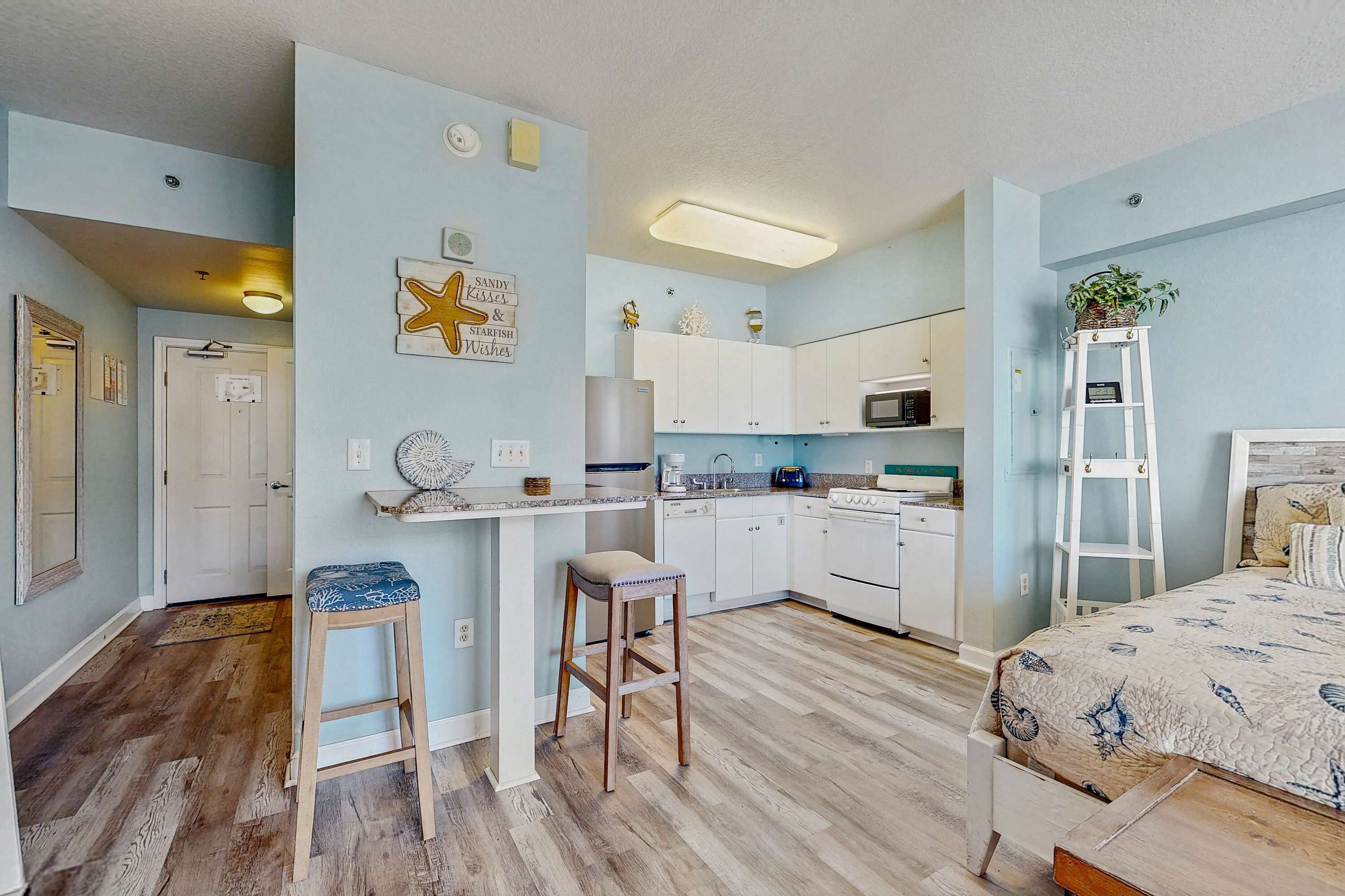 A compact kitchen with white cabinets, a microwave, and a small countertop. There is a wooden stool and a ladder with a plant on it.