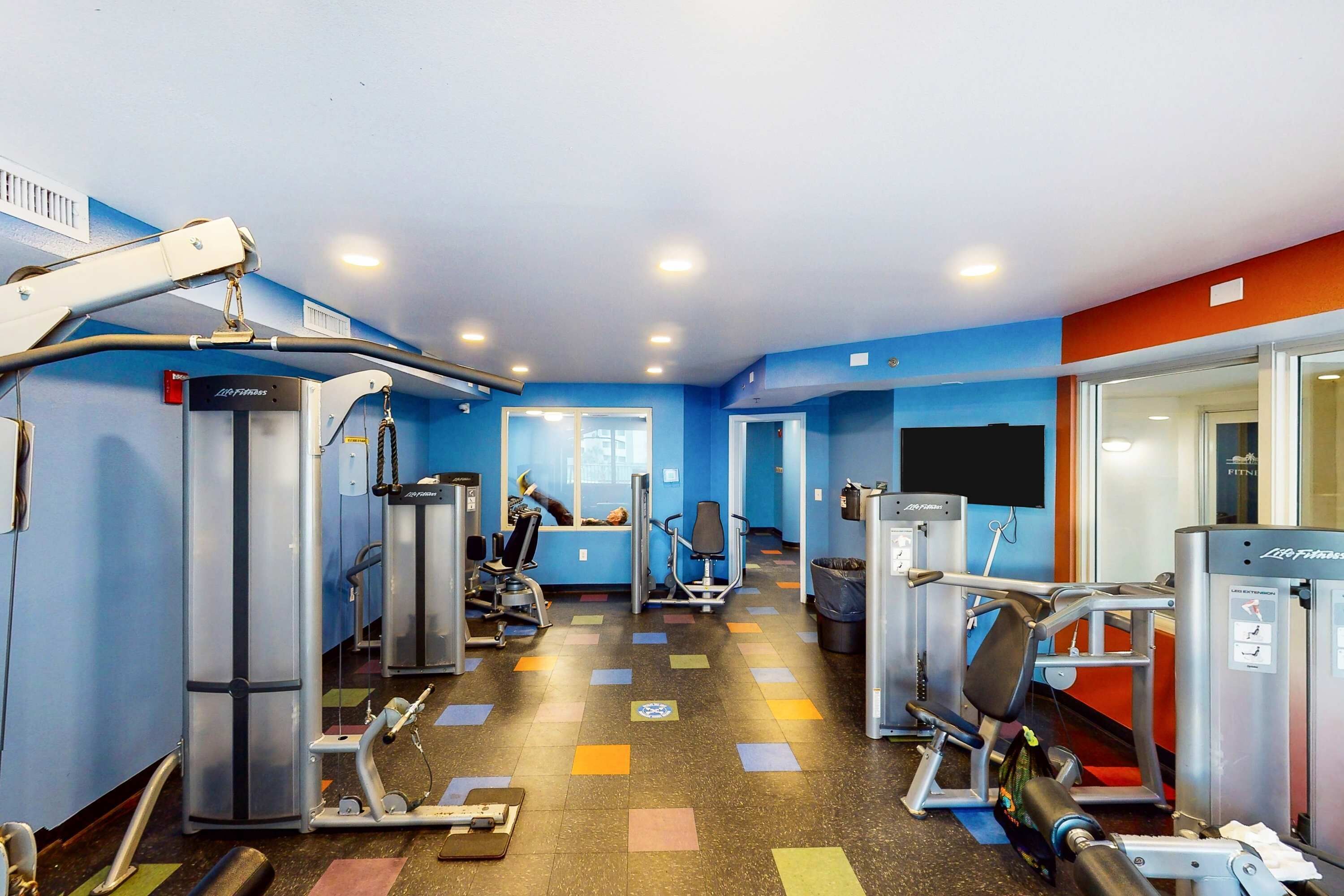 A gym with various exercise machines, a mirrored wall, and a colorful floor.