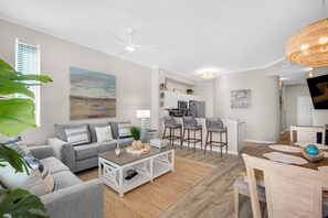 A modern living room with a gray sofa, a coffee table, and a dining area with a wooden table and chairs. - Beautiful 2nd-floor condo in Seagrove Beach with beach ambience & pickleball (Santa Rosa Beach)