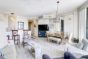 A spacious living area with a sofa, coffee table, and dining table set. A TV is mounted on the wall, and there are chairs and a bench. - Beautiful 2nd-floor condo in Seagrove Beach with beach ambience & pickleball (Santa Rosa Beach)
