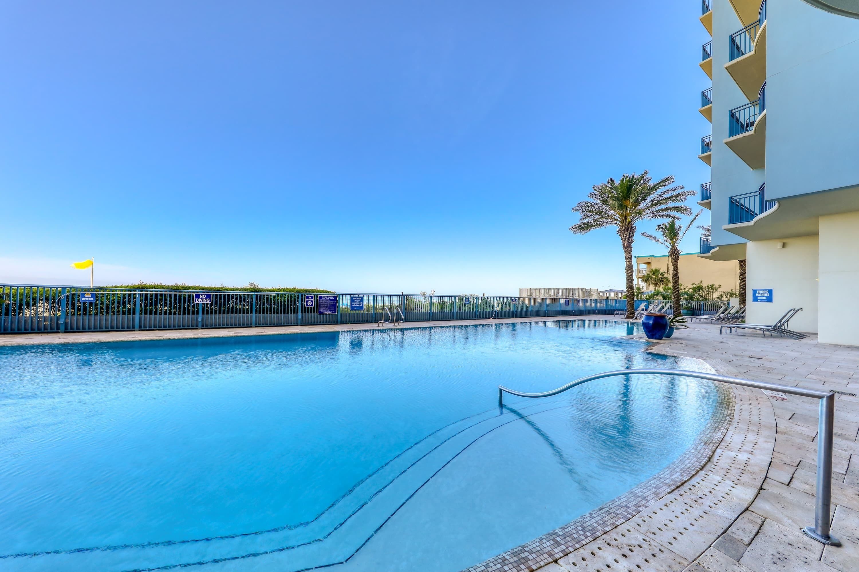 A clear blue swimming pool with a metal railing, palm trees, and a building with balconies.