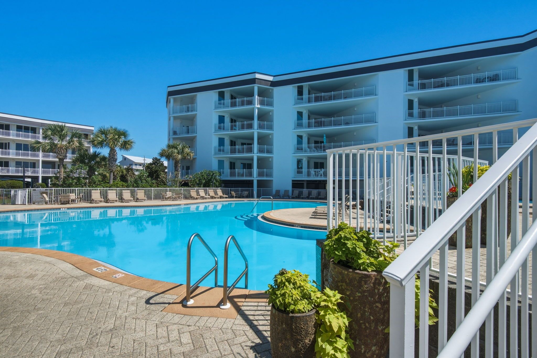 A swimming pool with a white fence, a brick walkway, and a multi-story building with balconies in the background.