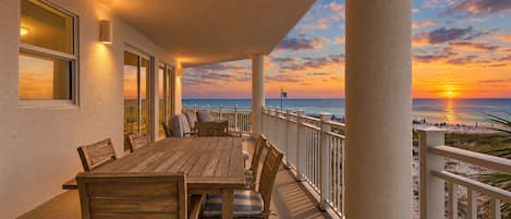 A wooden table and chairs on a balcony with a view of the sunset over the ocean.