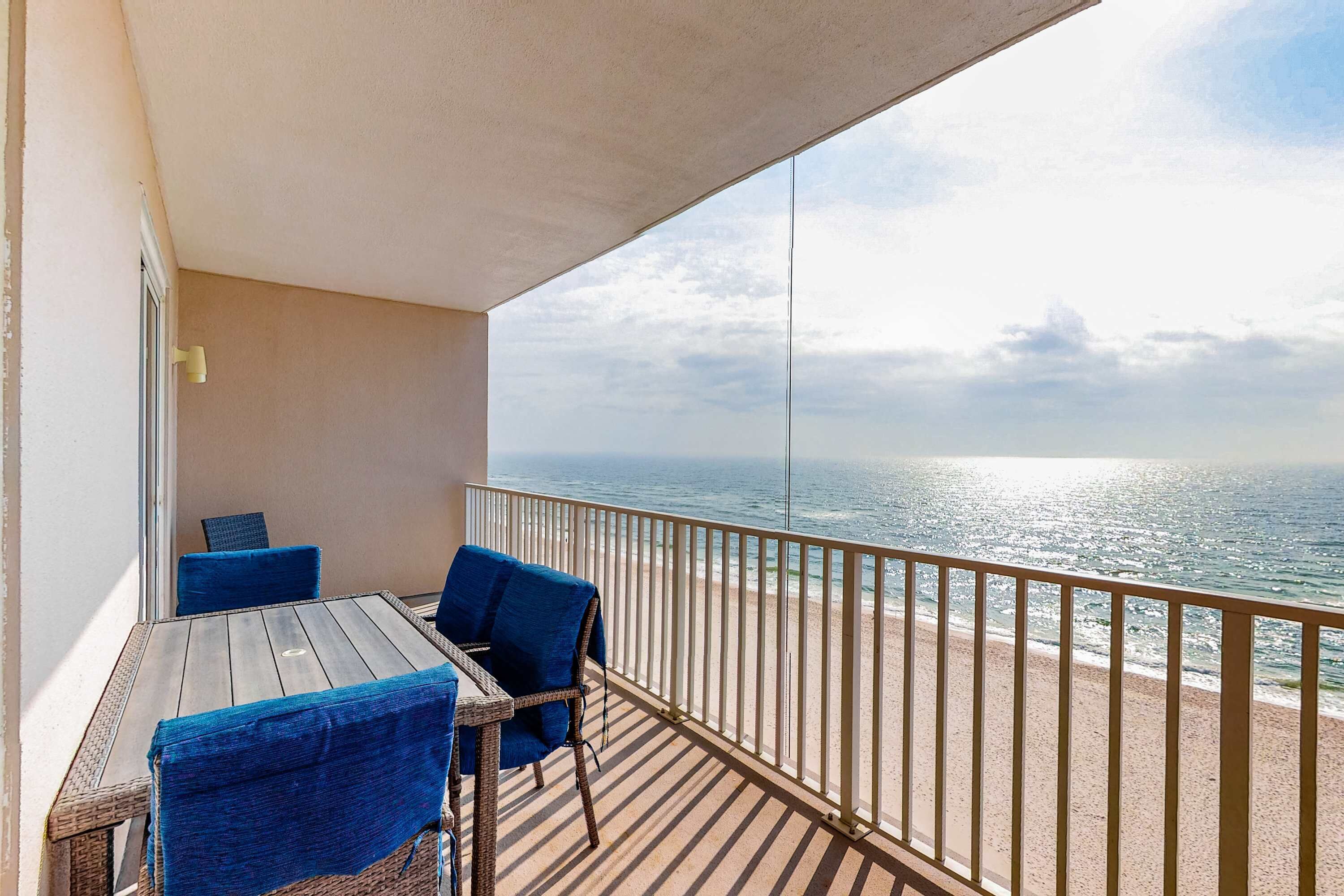 Un balcon avec une table en bois et des chaises bleues, donnant sur une plage et agrémenté d’une rambarde.