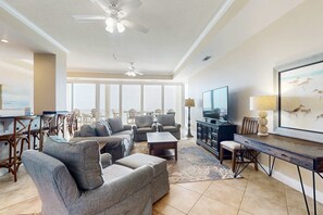 A spacious living room with a large window, a ceiling fan, and a flat-screen TV. - Beautiful, Gulf-front townhome with three water-view decks - near entertainment (Perdido Key)