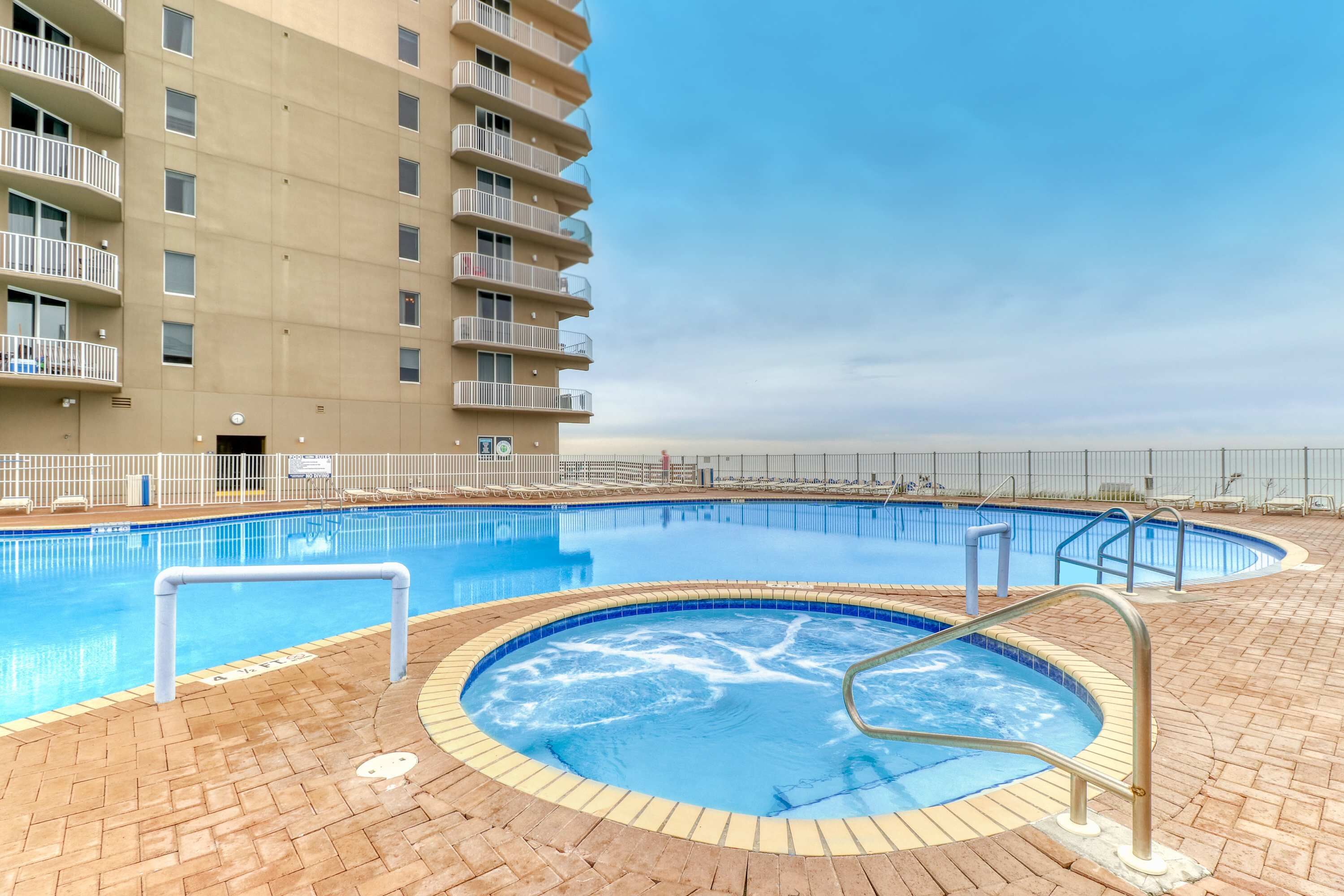 A swimming pool area with a hot tub, surrounded by a brick patio, and adjacent to a high-rise building with balconies.