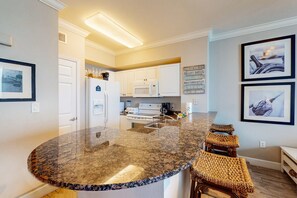 Una cocina moderna con armarios blancos, encimera de granito y microondas integrado. Hay dos cuadros enmarcados en la pared y un cartel con motivos náuticos.