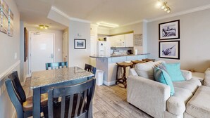 Une cuisine moderne avec des meubles blancs et un îlot central, un coin repas avec une table en granit et des chaises en bois, et un salon avec un canapé beige et des coussins turquoise.