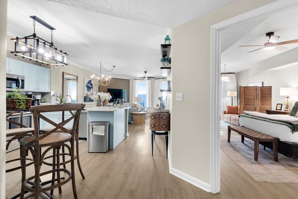Eine moderne Küche mit Kochinsel, ein Essbereich mit Holzkästen, ein Wohnzimmer mit Sofa und Deckenventilator sowie ein Flur, der zum Schlafzimmer führt.
