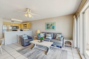 A modern living room with a grey sofa, a white coffee table, and a grey armchair. There is a kitchen area with wooden cabinets and a microwave. A ceiling fan is visible, and a painting hangs on the wall. - 2nd Floor Gulf Front Condo, Complimentary Beach Equipment, Nearby Shops & Dining (Destin)