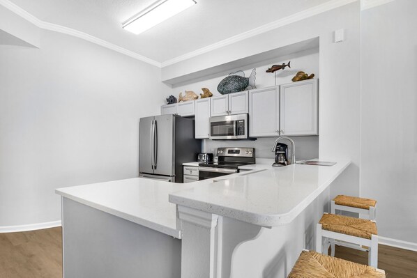 A modern kitchen with white cabinetry, a central island with stools, a refrigerator, a microwave, a toaster, and a coffee maker. - 4th-floor condo with beach ambience, lagoon pool, gym, tennis & pickleball (Santa Rosa Beach)