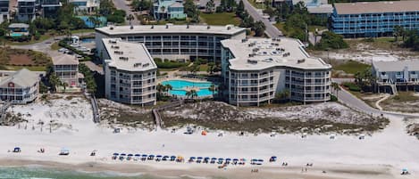 Un hotel frente a la playa con varias construcciones, piscina y una playa de arena con sombrillas.