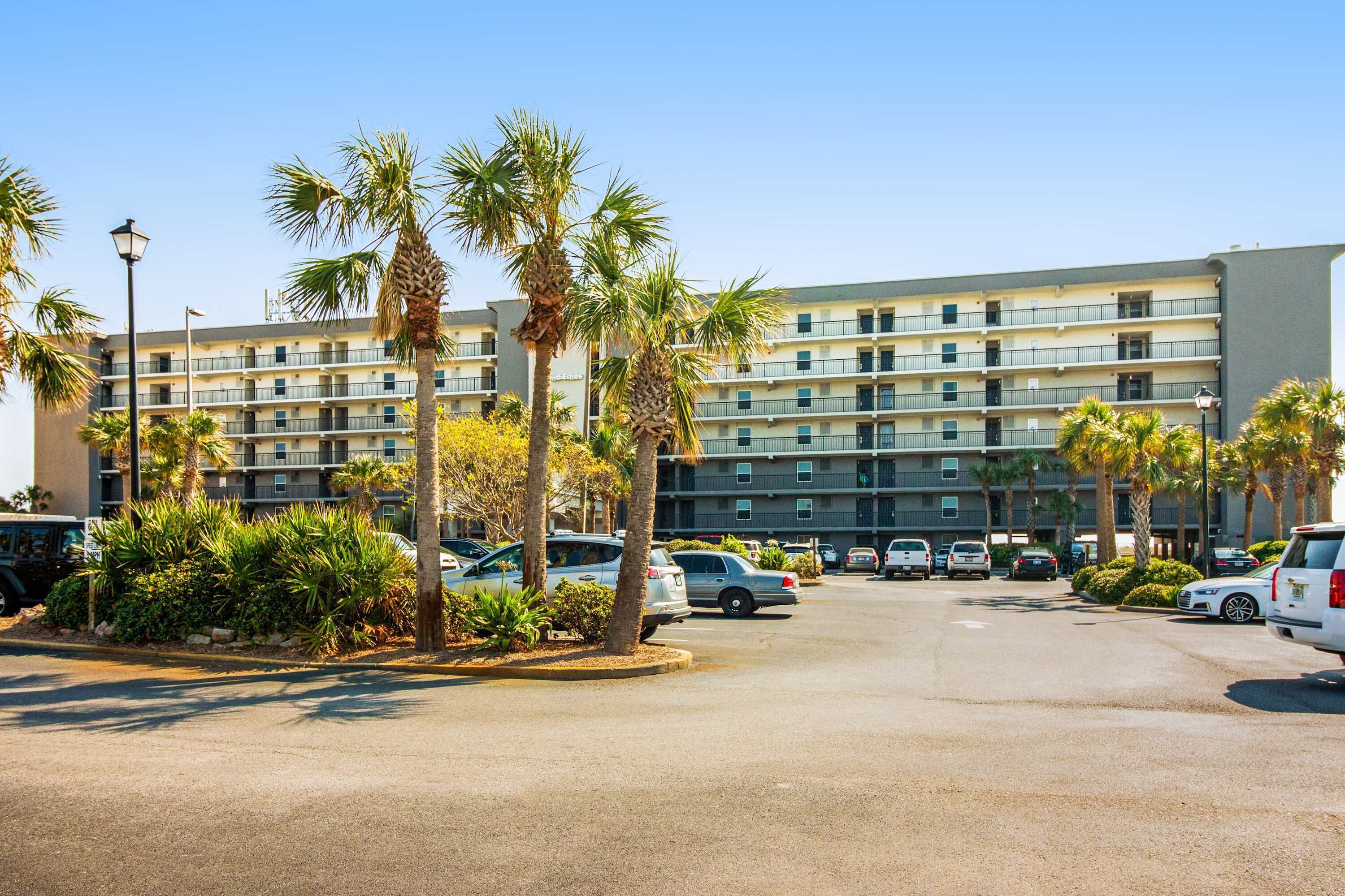 Een hotel met meerdere verdiepingen, een parkeerplaats, palmbomen en een helderblauwe lucht.
