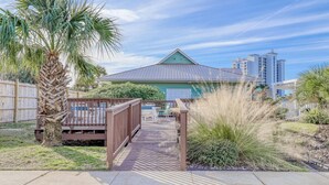 A wooden deck leading to a building with a green roof and white walls, surrounded by palm trees and tall grasses. - Ground-floor condo half block from the beach with patio, central AC, pool & WiFi (Destin)