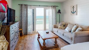 A living room with a wooden coffee table, a sofa, a TV on a dresser, and a view of the ocean through a sliding door. - Modern oceanfront studio with pool access, central AC, and beach service (Fort Walton Beach)