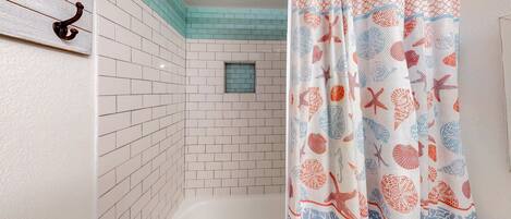 A bathroom with white tiled walls, a small window, and a shower curtain with a sea shell pattern.