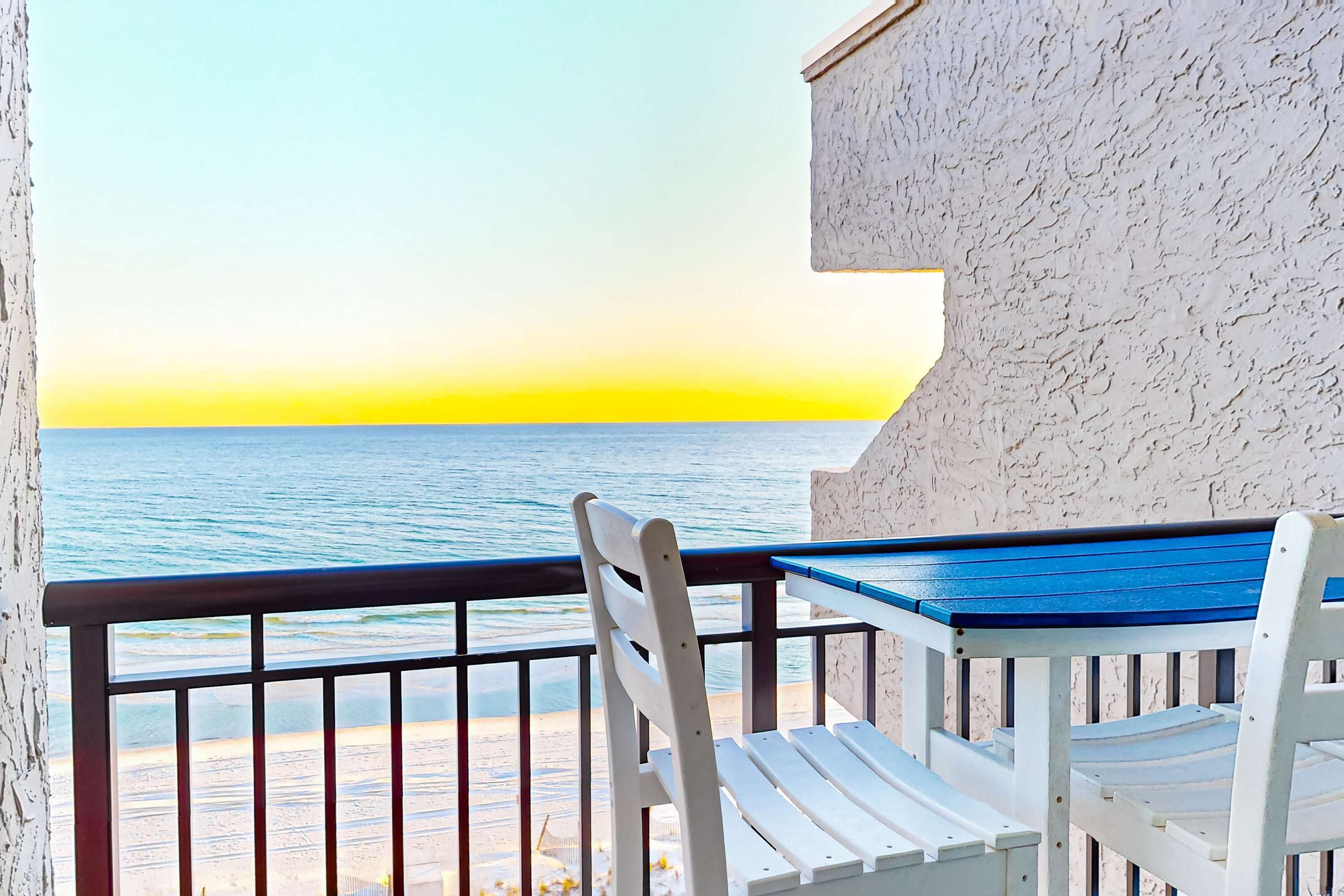 Uma varanda com uma cadeira branca e uma mesa azul, com vista para uma praia sob um céu claro e mar calmo.