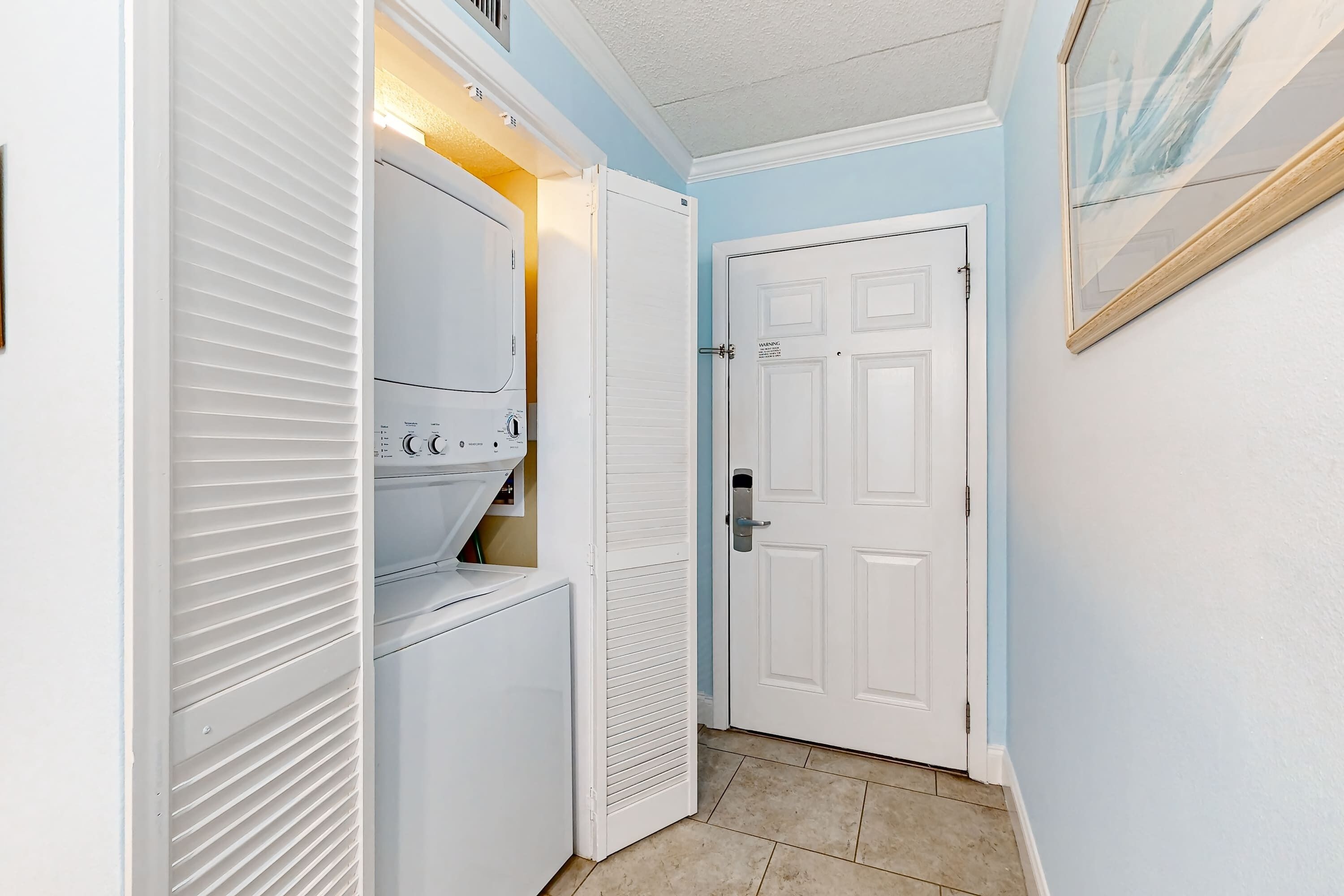 Un cuarto de lavandería con lavadoras y secadoras apiladas, una puerta blanca y un cuadro en la pared.