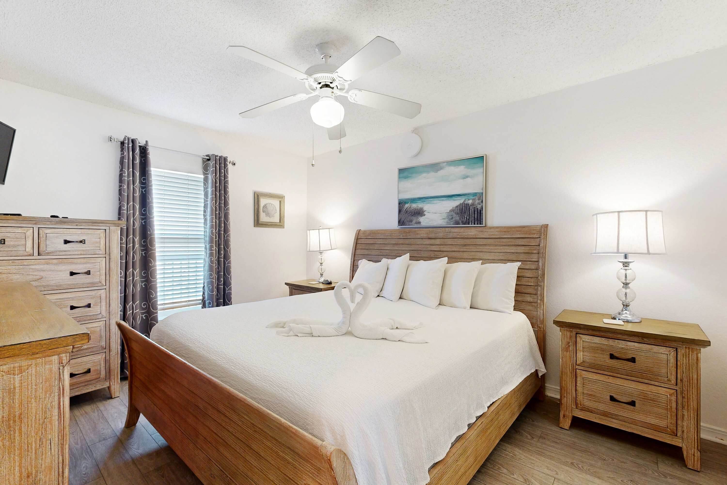 A bedroom with a wooden bed, a ceiling fan, bedside lamps, a nightstand, a dresser, and a window with curtains.