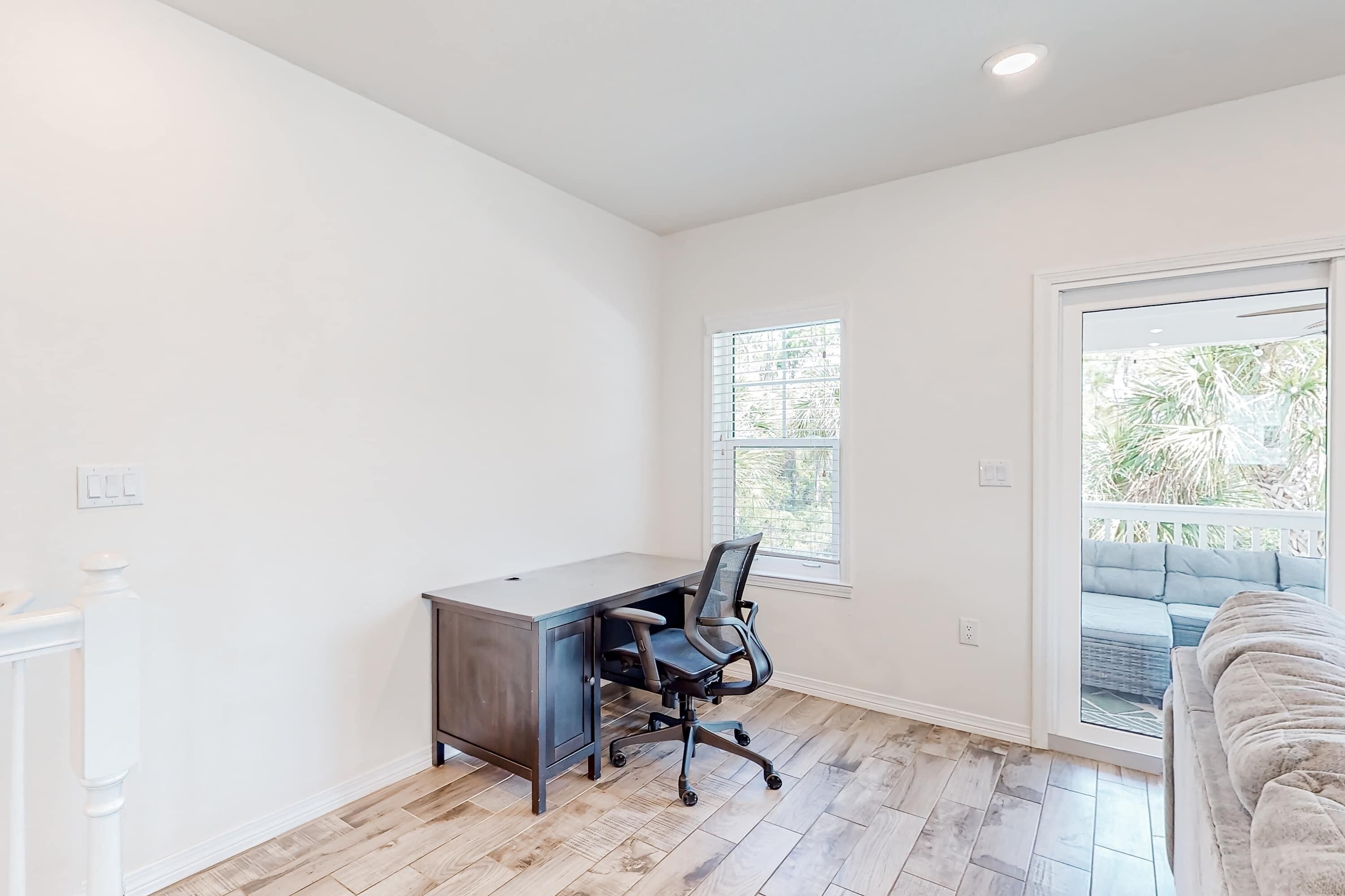 Une pièce spacieuse avec un plancher en bois, un bureau avec une chaise, une grande fenêtre et un balcon doté de mobilier d’extérieur.