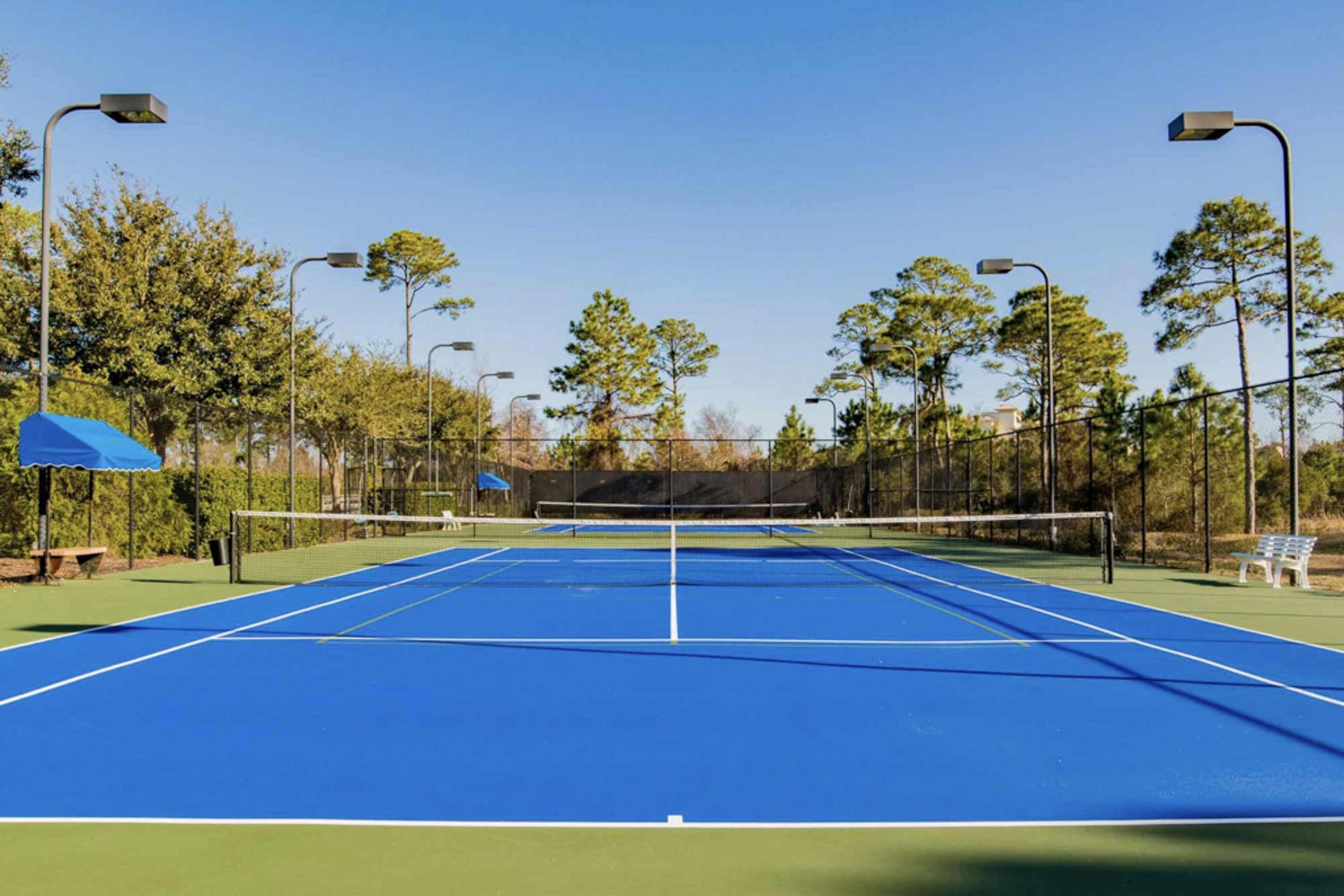 An outdoor tennis court with a net, surrounded by trees and street lamps.