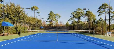 An outdoor tennis court with a net, surrounded by trees and street lamps.