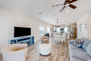 A spacious living room with a flat-screen TV, a ceiling fan, and a dining area with a kitchen in the background. - Dog-friendly townhouse with private beach, hot tub, pool, tennis, golf, gym, AC (Perdido Key)