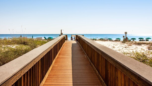 A wooden boardwalk leading to a beach with umbrellas and people in the distance. - Gulf-front condo with heated pool, fitness room & beach chairs - close to dining (Fort Walton Beach)
