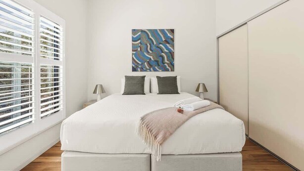 Une chambre à coucher comprenant un lit, deux tables de chevet, une fenêtre avec des stores et une œuvre d’art accrochée au mur.