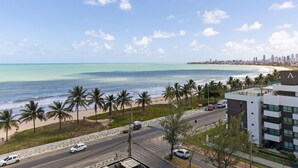 A coastal road with palm trees, a beach, and a city skyline in the background. - Mar Bello Plaza #207 por Carpediem (Cabedelo)