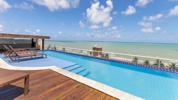 A pool with lounge chairs, a wooden deck, and a view of the ocean. - Mar Bello Plaza #207 por Carpediem (Cabedelo)