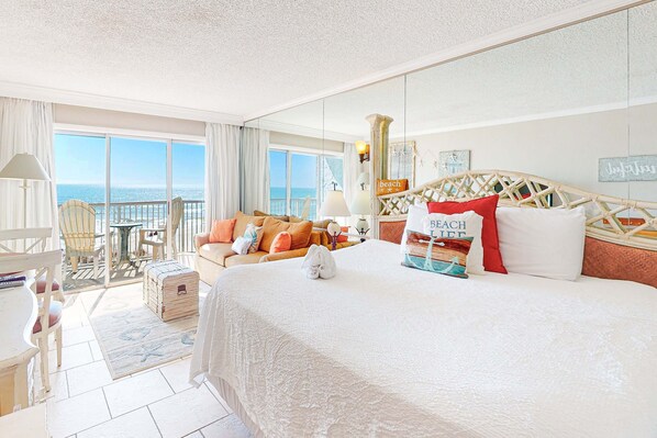 A hotel room with a large bed, a balcony with a view of the sea, a dining area, and a lounge with a view. - Attractive & Convenient Oceanfront Studio w Shared Pool, Gym, Free WiFi, AC & Balcony (Fort Walton Beach)