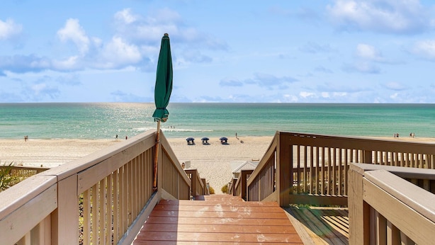 Een houten trap die naar een strand leidt, met een helder uitzicht op de oceaan en enkele parasols.