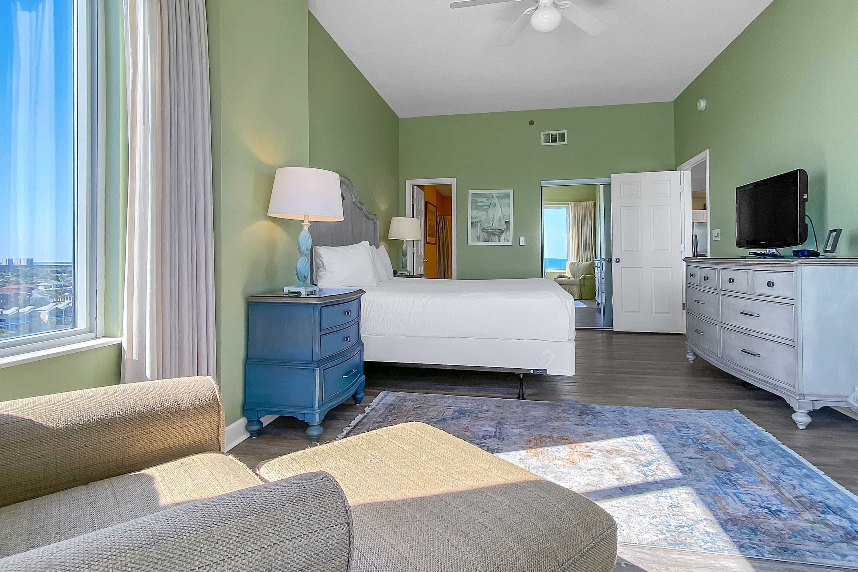 A hotel room with a bed, a blue dresser, a white dresser, a television, and a view of the city through the window.