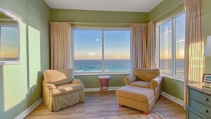 Um quarto com duas poltronas bege, uma pequena mesa redonda de vime e vista para o mar através de grandes janelas com cortinas bege.
