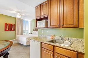 A hotel room with a kitchenette, a bed, a ceiling fan, and a large window. - Stylish oceanfront West Tower corner condo - 3 balconies, WiFi, & pool/hot tub (Panama City Beach)