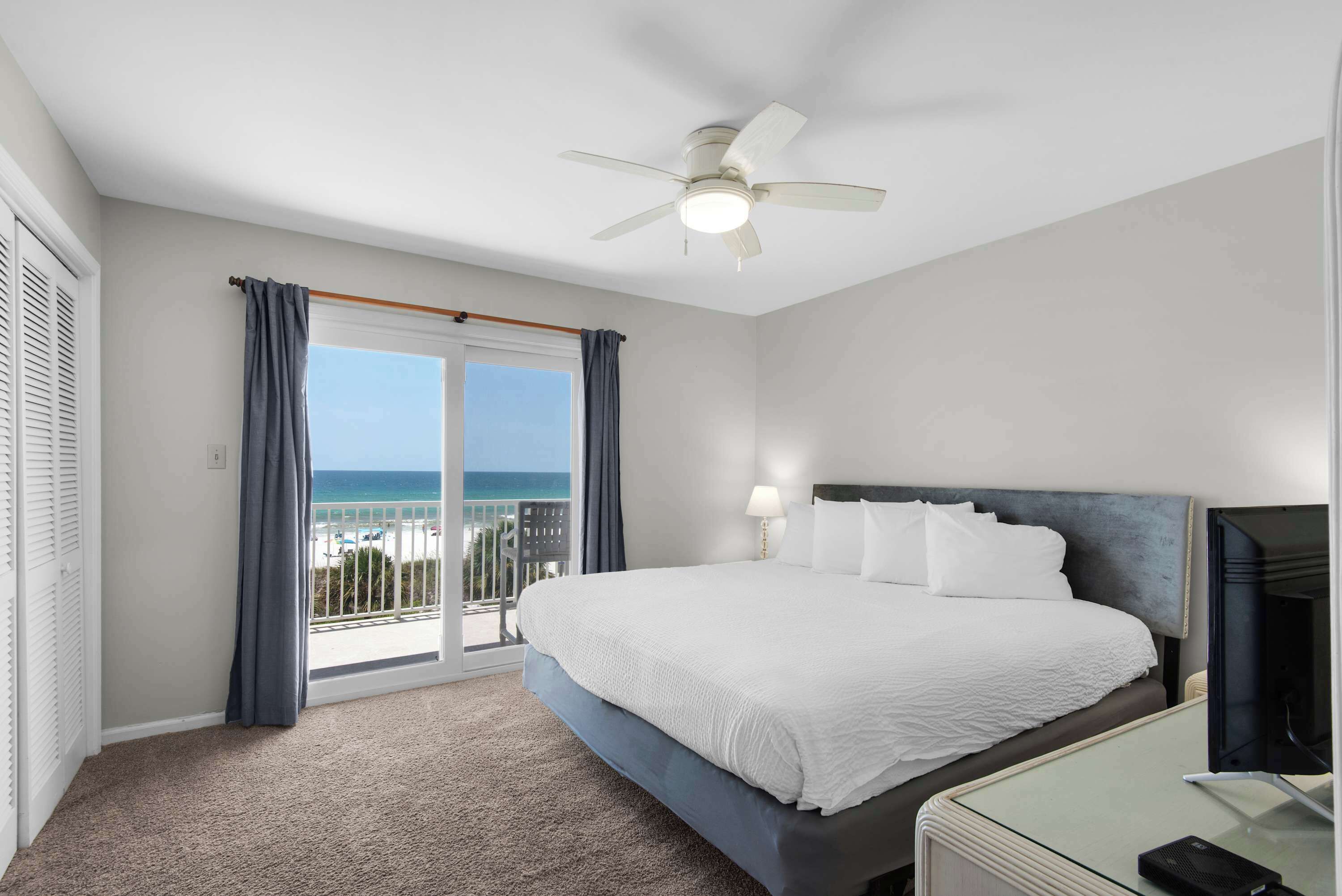 A hotel room with a bed, a desk, a television, a ceiling fan, and a view of the ocean.